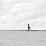 A lone runner traverses a vast, empty Brazilian beach under dramatic cloudy skies in black and white.